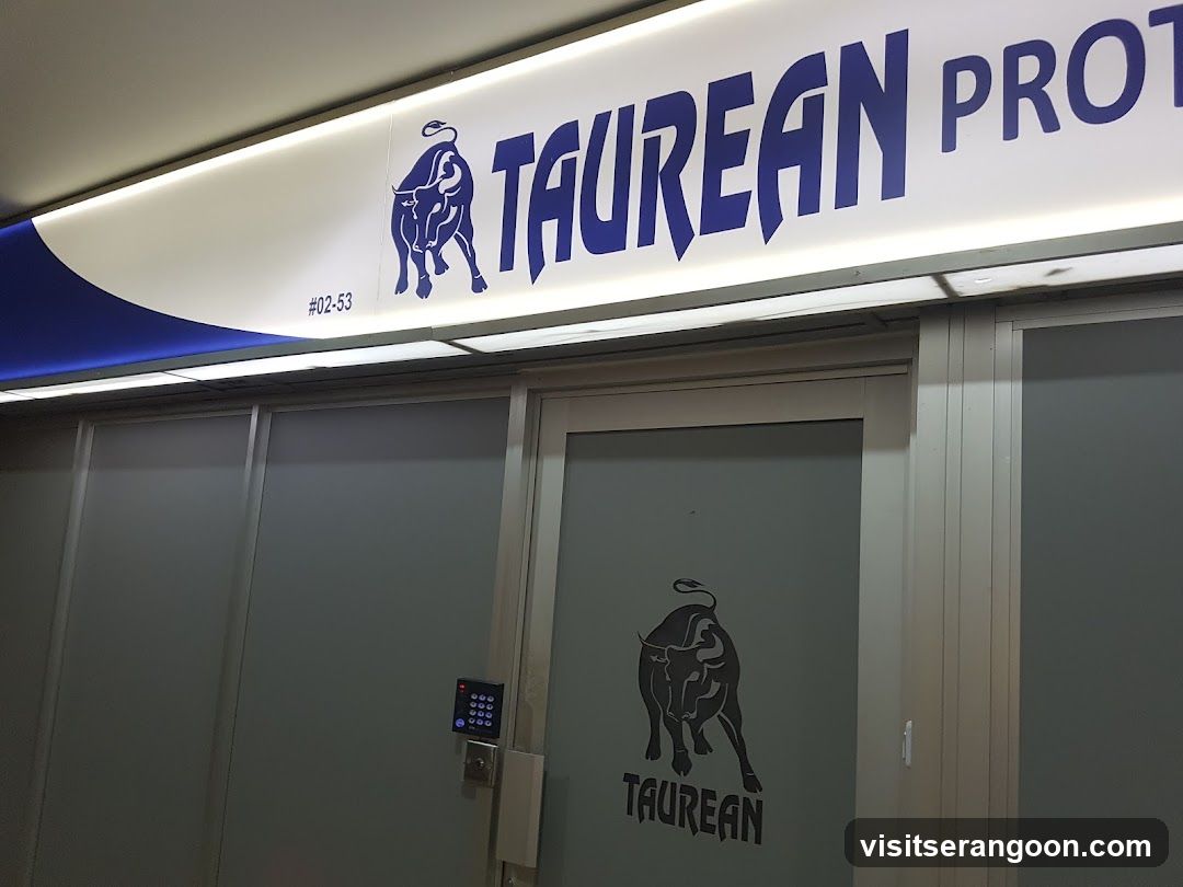 A view of a commercial storefront featuring a large, illuminated overhead sign with the text 'TAUREAN PROT' in bold blue letters next to a graphic of a charging bull. The unit number #02-53 is visible on the sign. Below, there is a glass door with a matching bull logo and the word 'TAUREAN' printed on the glass, along with a digital keypad access system mounted on the wall.