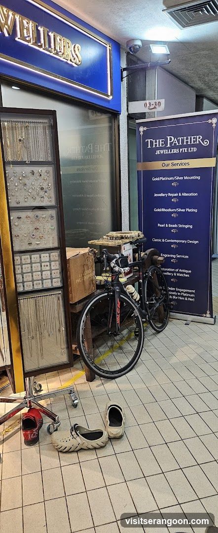 A view of a jewelry store entrance with a blue sign reading 'JEWELLERS' and a vertical banner listing services like gold mounting and repair. A bicycle is parked in front of the glass door, and shoes are on the tiled floor.