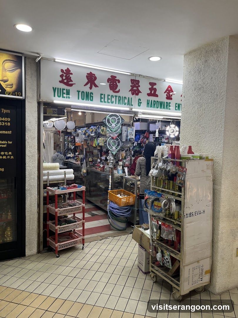 A view of a hardware and electrical shop entrance located in a tiled corridor. The storefront features a large white sign with red Chinese characters and green English text reading 'YUEN TONG ELECTRICAL & HARDWARE ENT'. The shop is open, revealing shelves stocked with various goods including light fixtures, rolls of material, and tools. Outside the entrance, there are metal shelving units displaying products like spray cans and packaged items, along with a red rolling cart and stacked boxes.