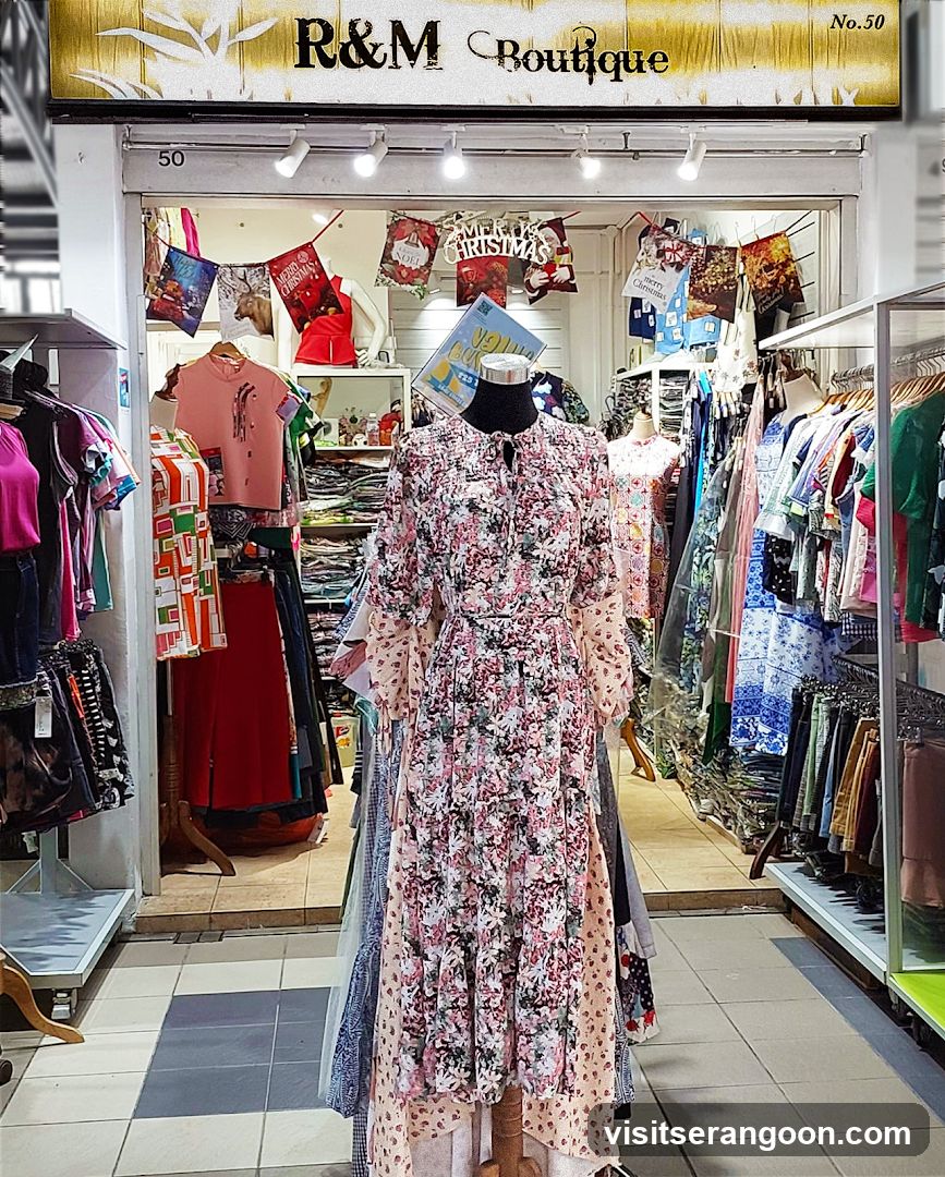 A view of the entrance to a clothing boutique named R&M Boutique. The storefront features a large sign with gold and white abstract patterns. Inside, racks of colorful clothing are visible, along with a central mannequin wearing a long floral dress. Christmas-themed decorations hang from the ceiling.