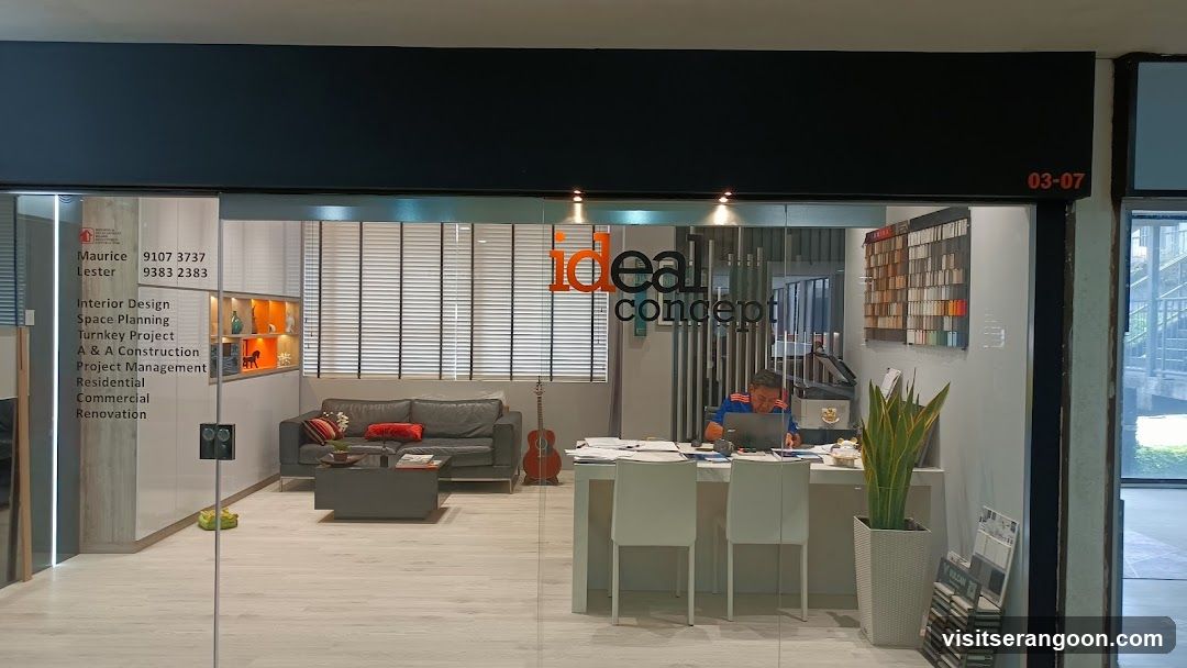 A glass-fronted office or showroom with a black fascia board displaying the unit number 03-07. The glass facade features the company name 'ideal concept' in orange and black lettering, along with a list of services including Interior Design, Space Planning, and Construction. Inside, a man works at a white desk, and a lounge area with a grey sofa and guitar is visible.