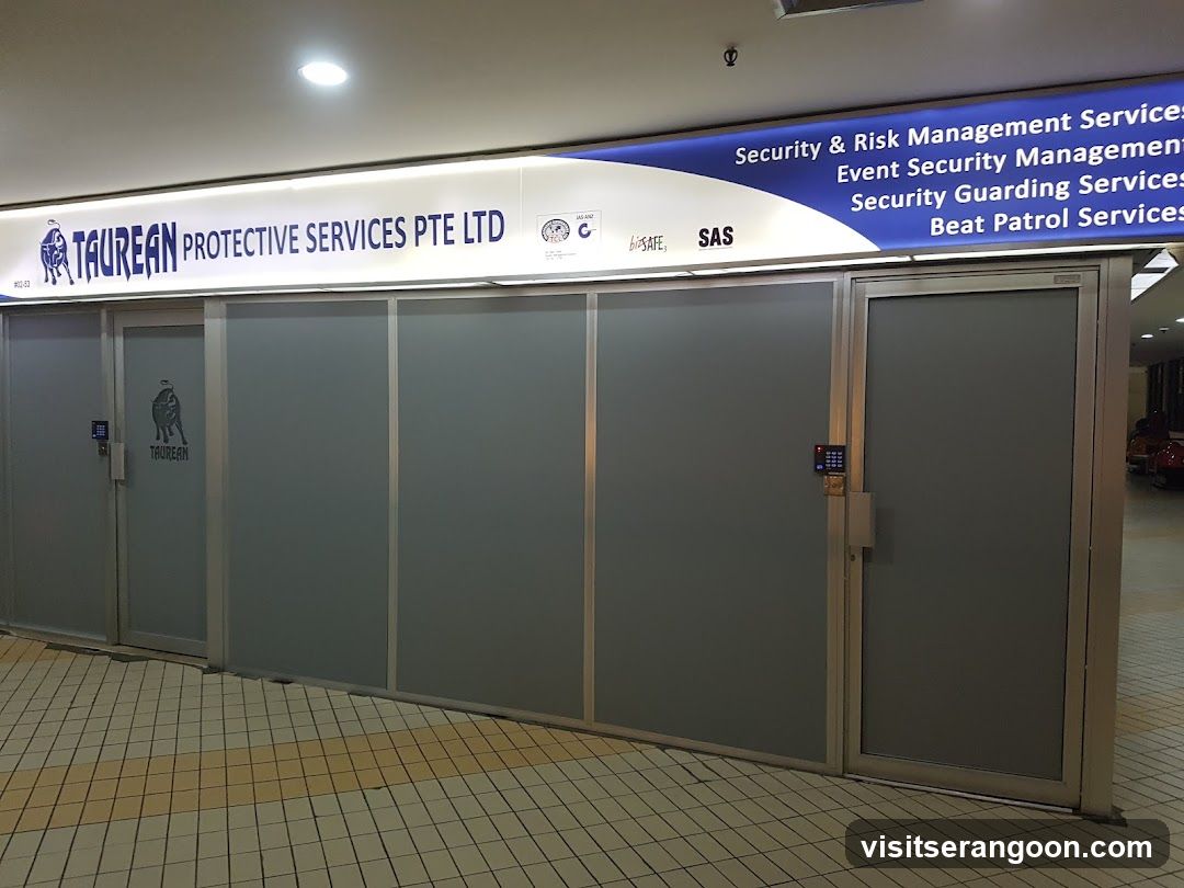 A view of a commercial storefront featuring a large overhead sign for 'TAUREAN PROTECTIVE SERVICES PTE LTD'. The sign lists services including Security & Risk Management, Event Security Management, Security Guarding Services, and Beat Patrol Services. The entrance consists of frosted glass sliding doors with electronic access control keypads mounted on the frames.