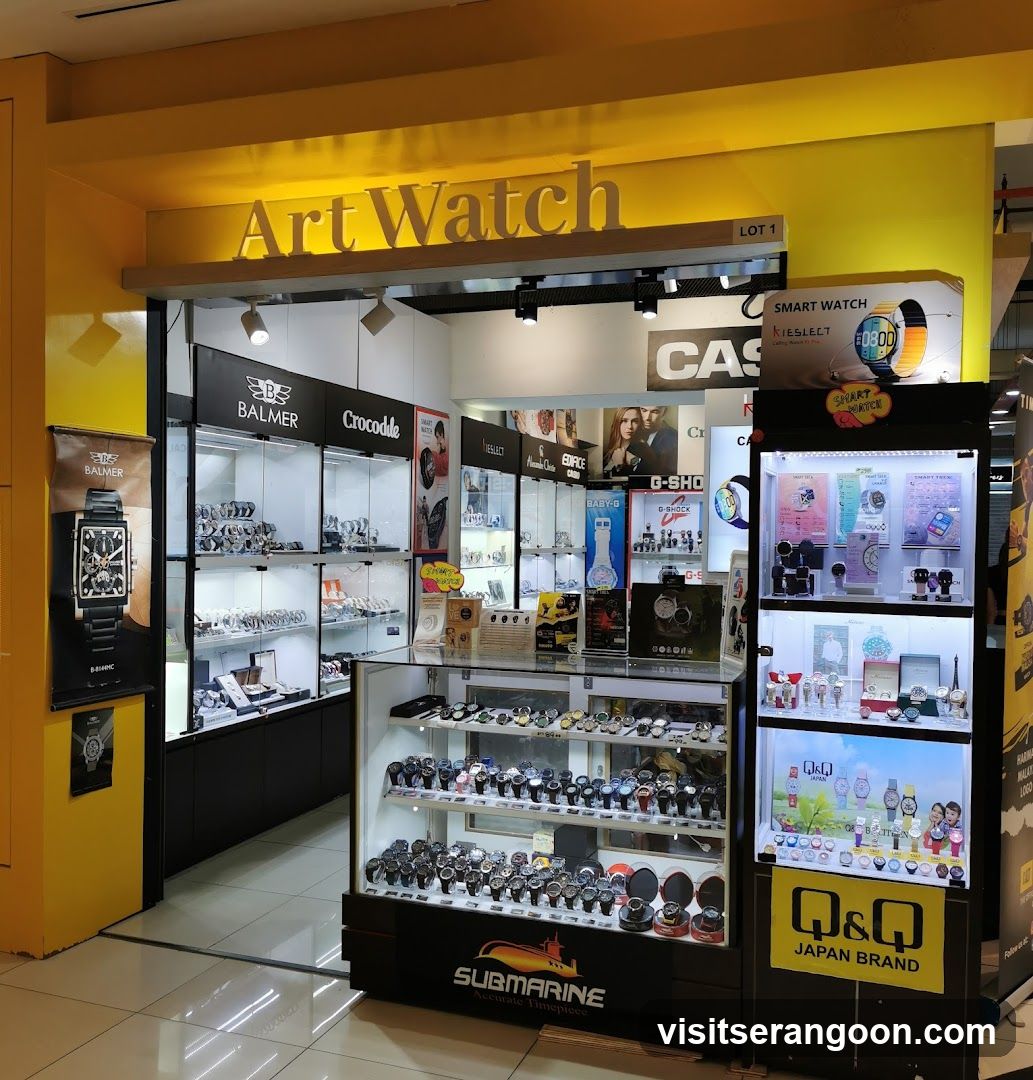 A brightly lit retail kiosk with a yellow facade and the sign 'Art Watch' above the entrance. The store displays numerous watches in glass cases, featuring brands like Balmer, Crocodile, Casio, and Q&Q. A large poster on the left shows a specific watch model, while shelves on the right display colorful smartwatches and other accessories.