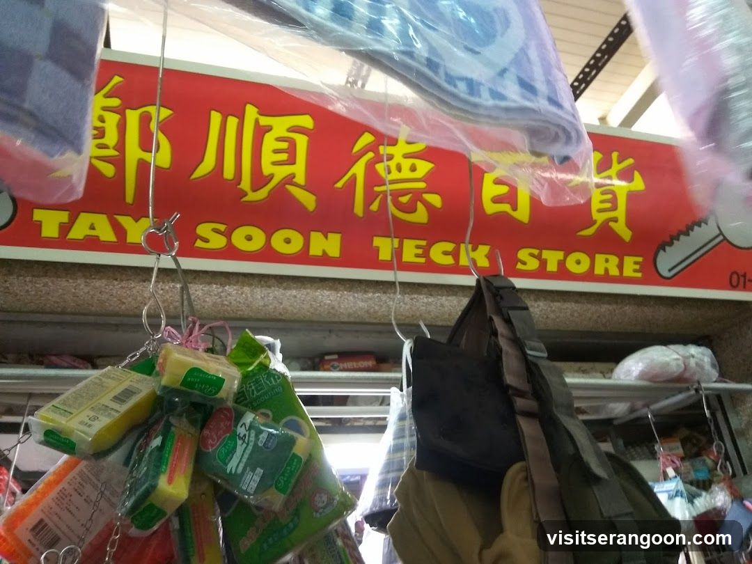 A close-up view of a storefront featuring a prominent red sign with yellow Chinese characters and the English text 'TAY SOON TECK STORE'. Below the sign, various merchandise is displayed, including packaged sponges and cleaning supplies hanging from hooks, alongside a dark-colored bag.