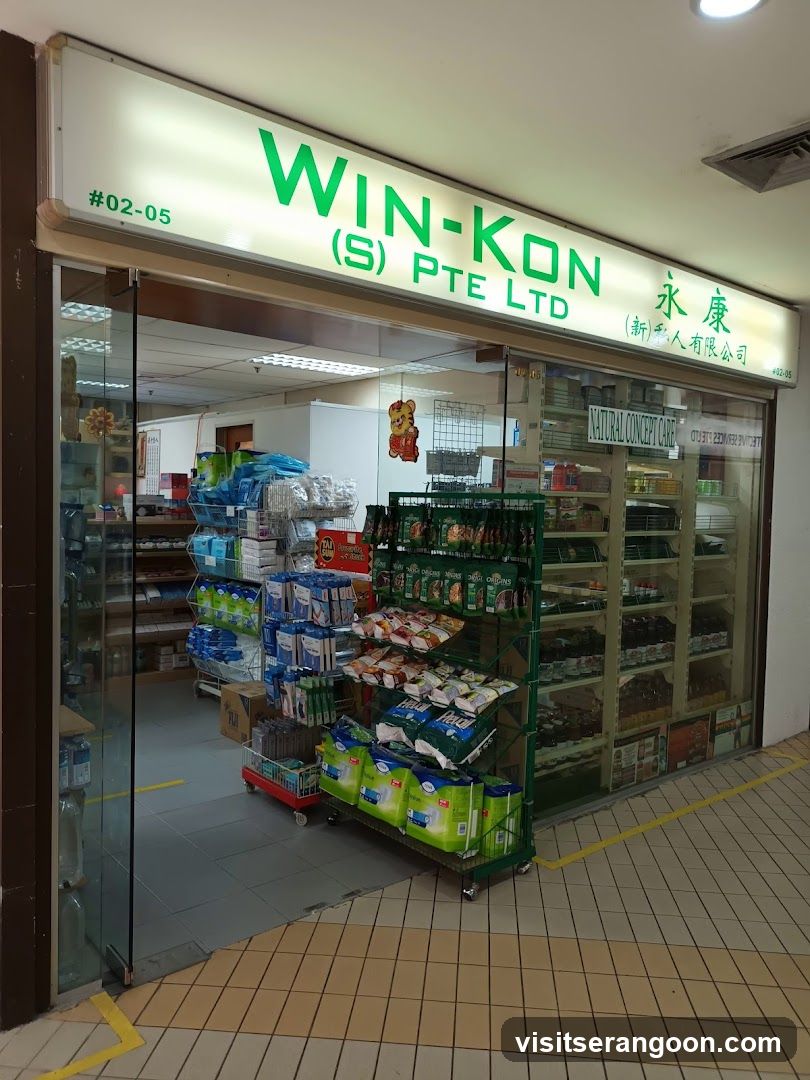 A brightly lit storefront with a large white sign displaying 'WIN-KON (S) PTE LTD' in green letters, alongside Chinese characters. The unit number #02-05 is visible on the left. The entrance features glass sliding doors and large glass windows, revealing shelves stocked with various packaged goods, including snacks and household items, inside the store.