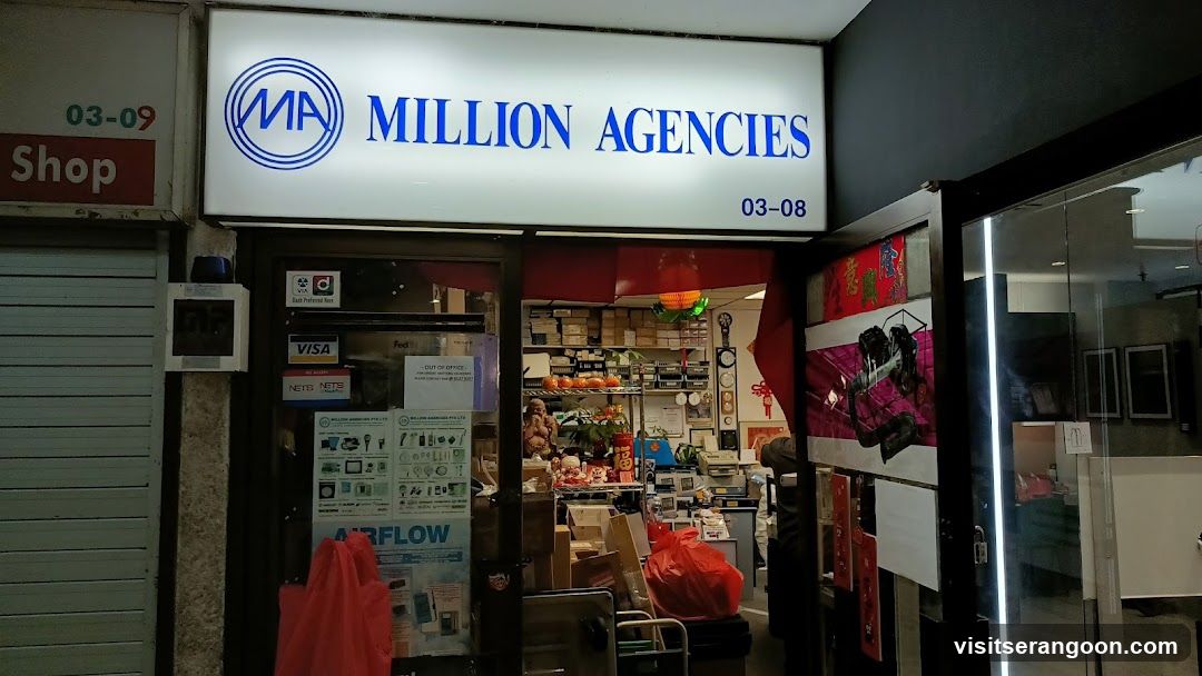 A brightly lit storefront with a large white sign reading 'MILLION AGENCIES' in blue text, flanked by unit numbers 03-09 and 03-08. The entrance features glass doors displaying various stickers, including payment logos like Visa and NetS, and a poster advertising 'AIRFLOW'. Inside, shelves are stocked with boxes and merchandise, and red decorations hang from the ceiling.