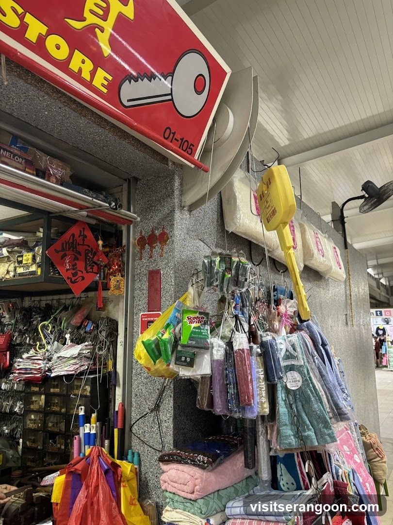 A view of a hardware store storefront with a red sign reading 'STORE' and a key graphic. The entrance is open, revealing shelves of goods inside. Outside, various items like towels, mops, and bags are displayed for sale.