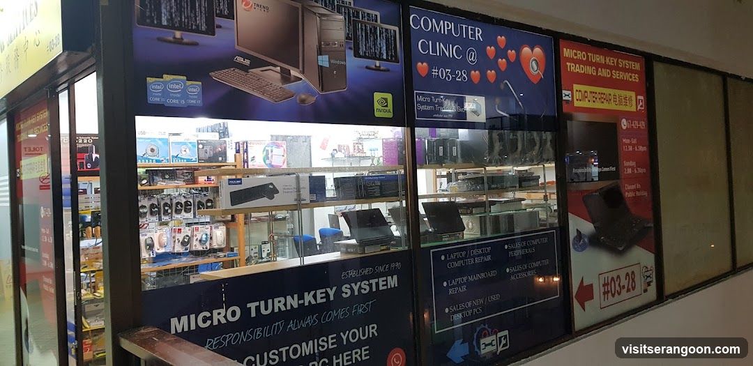 A storefront window display for a computer repair shop. The glass is covered with large decals advertising services like laptop and desktop repair, sales of new/used computers, and accessories. A prominent sign reads 'COMPUTER CLINIC @ #03-28'. Inside, shelves are stocked with boxed computer components, peripherals, and a laptop on a counter.