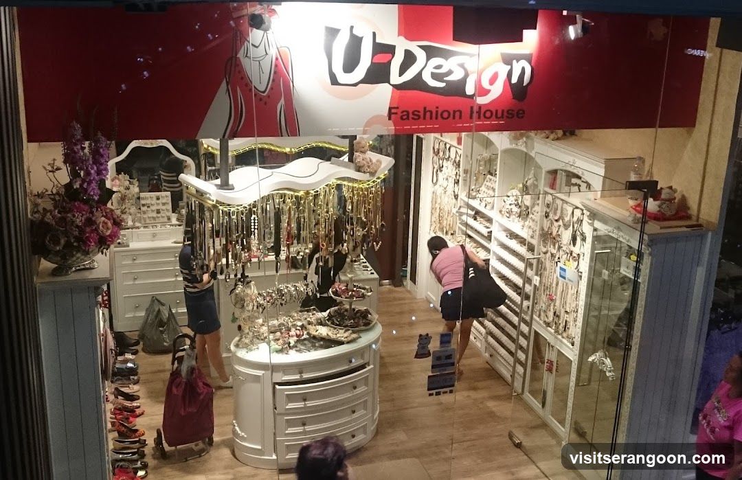 A view through a glass storefront window of a jewelry and accessories shop. A large red and white sign above reads 'U-Design Fashion House'. Inside, the store is brightly lit with white furniture, including a central display island and wall-mounted glass cabinets filled with necklaces and other items. Several customers are browsing the merchandise.