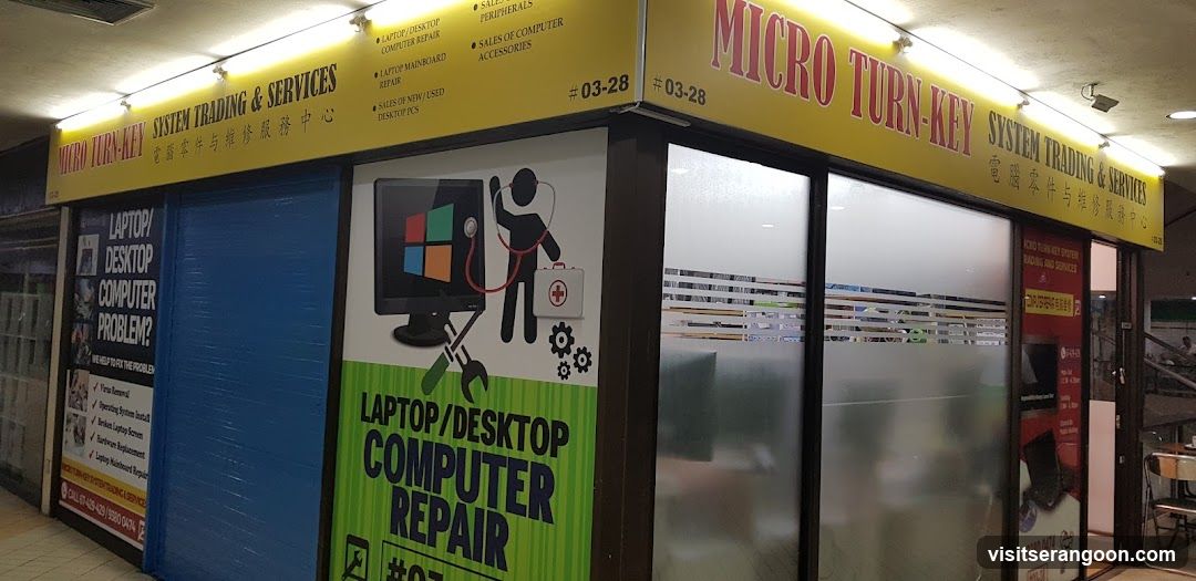 A storefront for a business named 'Micro Turn-Key' located in a shopping center. The yellow signage lists services like computer repair and sales of accessories. A large window display features a graphic of a doctor with a stethoscope next to a computer, advertising laptop and desktop repair services.