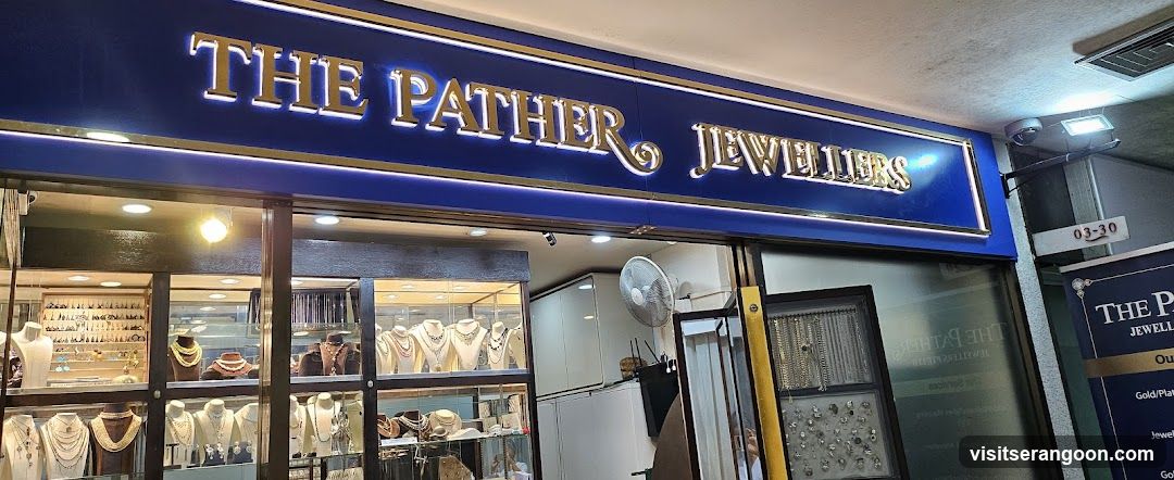 A photograph of a jewelry store exterior featuring a large blue sign with gold lettering reading 'THE PATHER JEWELLERS'. The storefront has large glass windows displaying various necklaces and jewelry on white busts inside. A white fan is visible inside near the entrance, and a small sign with the number 03-20 is attached to the wall on the right.