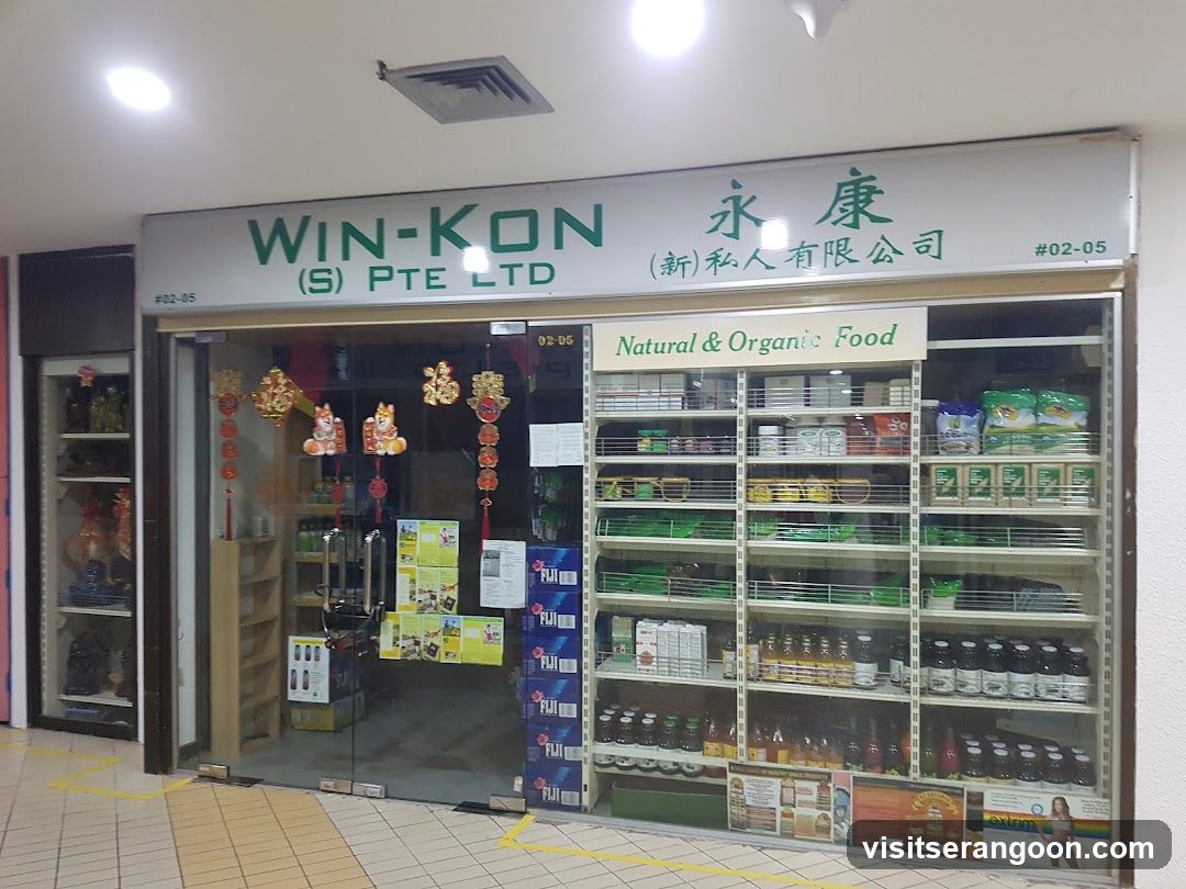 A storefront with a large white sign reading 'WIN-KON (S) PTE LTD' and Chinese characters. The entrance features glass doors decorated with hanging red and gold festive ornaments. To the right, a large display window showcases shelves stocked with various bottled beverages and packaged goods under a sign that reads 'Natural & Organic Food'.