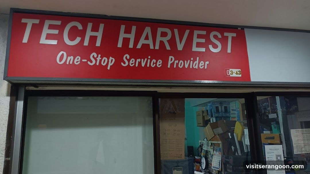 A red storefront sign with white text reading 'TECH HARVEST' and 'One-Stop Service Provider'. The shop number '03-43' is visible in the bottom right corner of the sign. Below the sign, glass windows reveal a cluttered interior with cardboard boxes and various items.
