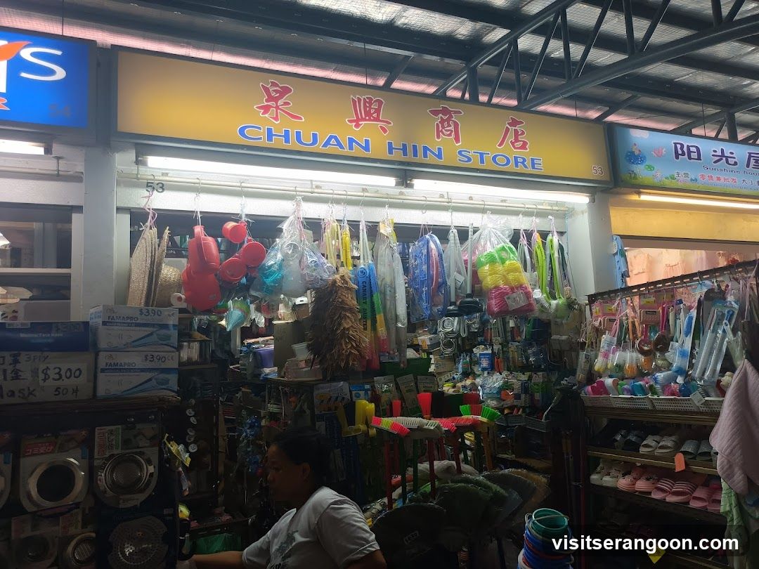 A brightly lit storefront with a large yellow sign displaying 'CHUAN HIN STORE' in red and blue text alongside Chinese characters. The shop is open, revealing shelves and hanging racks filled with various household goods, including colorful plastic buckets, brooms, mops, and cleaning supplies. A person is seated inside near the entrance.