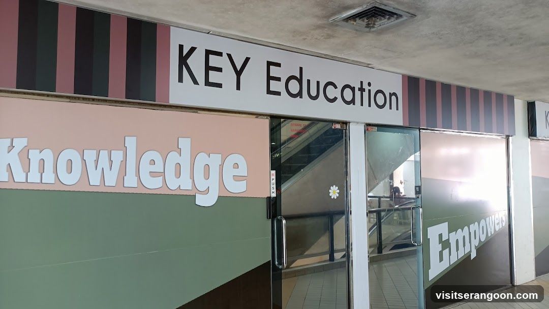 A storefront entrance for Key Education featuring a white sign with black text reading 'KEY Education'. The glass doors have large white lettering on them, with 'Knowledge' on the left panel and 'Empowers' on the right. The facade includes pink and black vertical stripes above the signage.