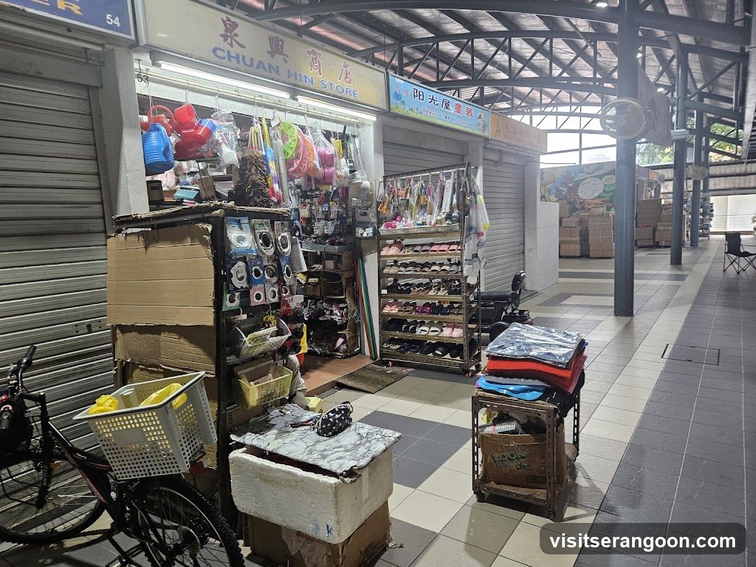 A view of a covered market aisle featuring a shop with open metal shutters. The storefront displays merchandise including colorful plastic buckets, hanging bags, and a multi-tiered shoe rack. A bicycle with a basket is parked in the foreground, and stacks of folded clothing sit on a wooden crate nearby. The shop has a sign with Chinese characters and English text reading 'CHUAN HIN STORE'.