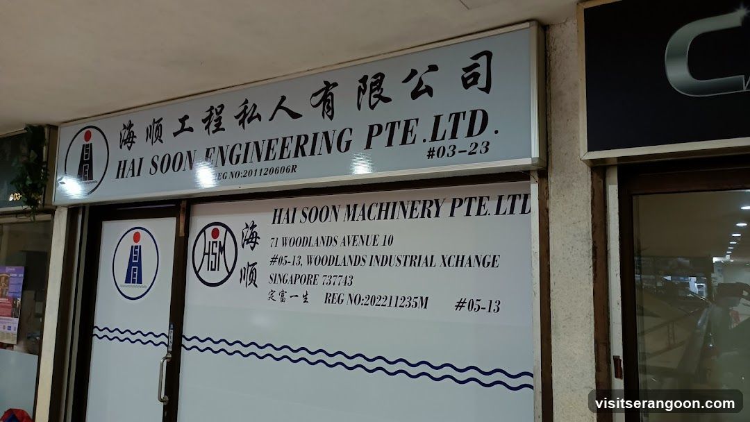 A photograph of a commercial storefront featuring two large white signs with black text. The top sign displays the company name 'HAI SOON ENGINEERING PTE. LTD.' along with a logo and registration number. Below it, a second sign reads 'HAI SOON MACHINERY PTE. LTD.' with an address in Woodlands, Singapore. The entrance consists of glass doors and windows with blue wave patterns near the bottom.