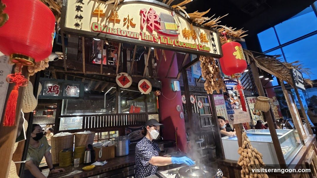 A street food stall with a large wooden sign reading 'Chinatown Tan's Tofu Coconut Cake' in English and Chinese characters. The stall features rustic decor including hanging red lanterns, dried corn stalks, and woven baskets. Staff members wearing masks and gloves are visible working behind the counter, preparing food in large metal pots.