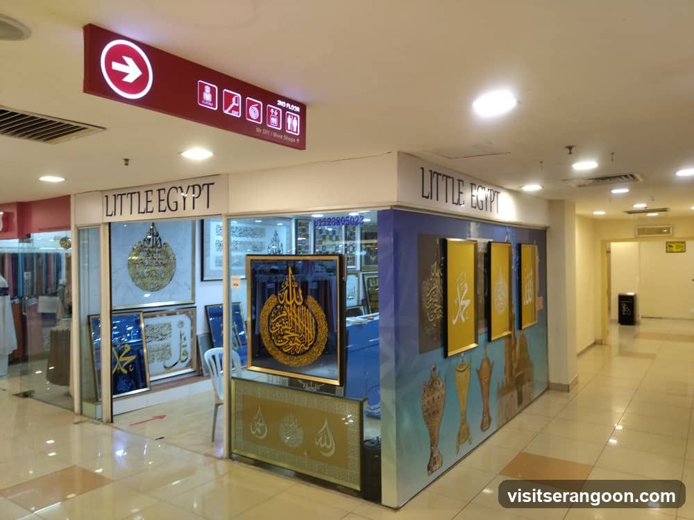 A storefront located in a shopping mall corridor with the name 'LITTLE EGYPT' displayed on the facade. The shop features glass windows displaying framed Islamic calligraphy art, including gold and blue pieces. A red overhead directional sign with a white arrow points towards the store.