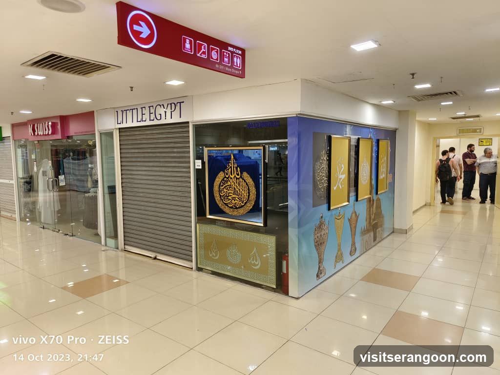 A view of a closed storefront inside a shopping mall. The store is named 'LITTLE EGYPT' in large black letters above a closed metal shutter. The facade features a large glass window displaying framed Islamic calligraphy art, including a prominent gold and blue piece. To the left is another store with a red sign reading 'K SWISS'. A red overhead directional sign hangs from the ceiling. Several people are standing in the hallway to the right.