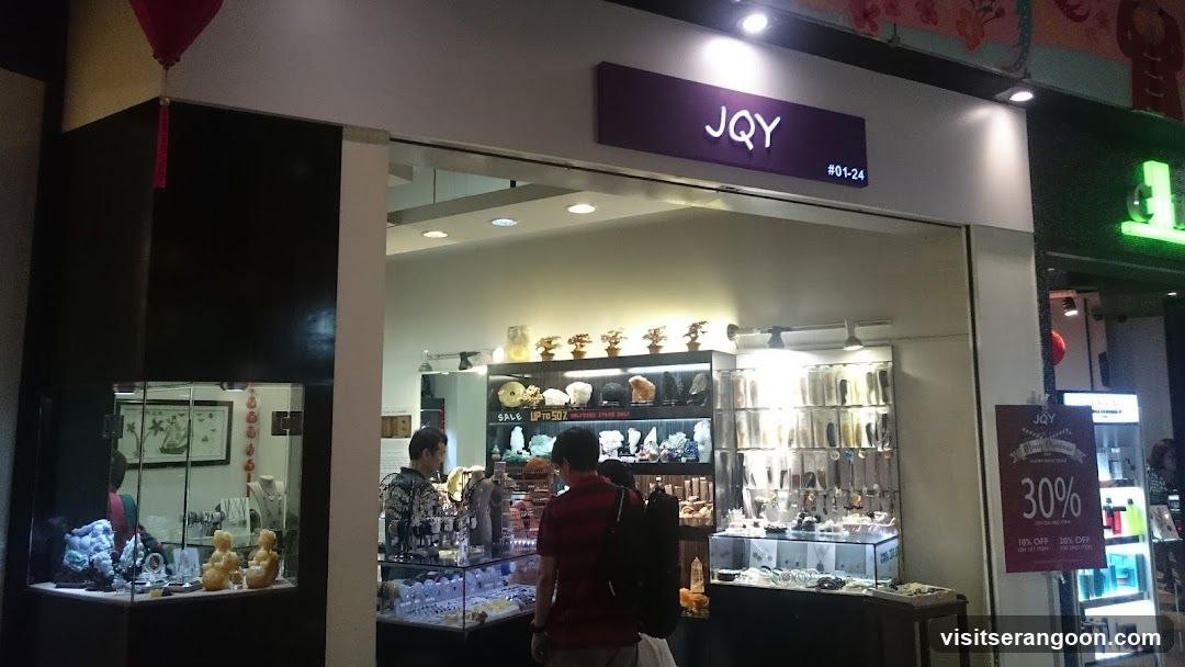 A view of a retail store entrance with a purple sign reading 'JQY' and unit number '#01-24'. Inside, glass display cases are illuminated, showcasing various items including what appear to be jade figurines and jewelry. A customer in a red shirt is browsing near the counter. A promotional sign on the right advertises '30% OFF'.