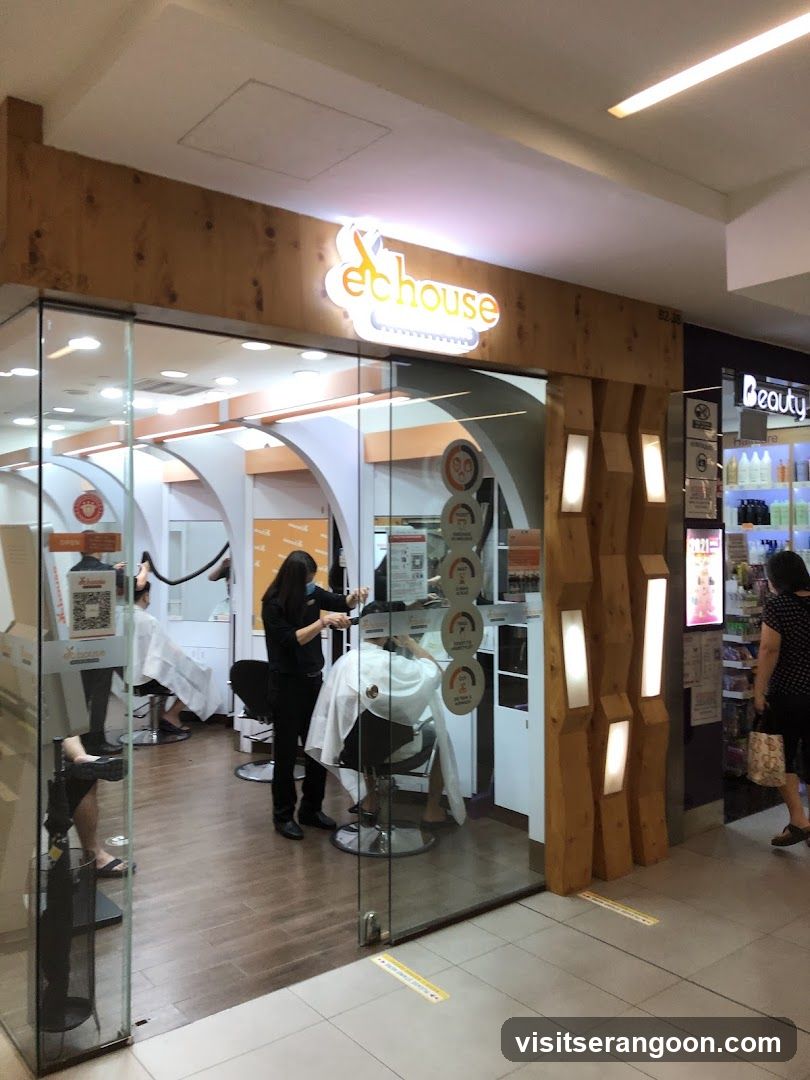 A view of the Echo House salon storefront located within a shopping mall. The entrance features a wooden facade with a backlit 'echo house' sign. Inside, through glass doors, stylists are seen working on clients in salon chairs. To the right, a neighboring store labeled 'Beauty' displays shelves of products.