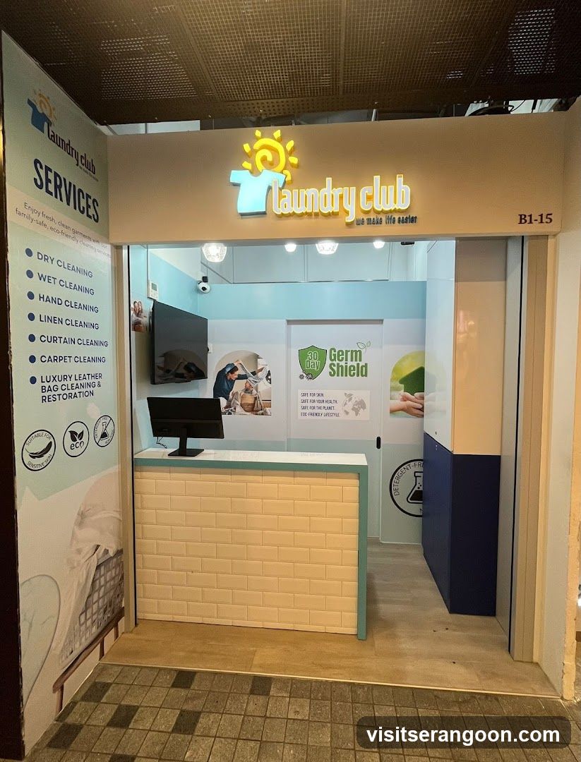 A view of the exterior entrance to a laundry service shop named 'Laundry club'. The storefront features a large illuminated sign with a sun and t-shirt logo above the entrance. To the left of the entrance, a vertical banner lists services including dry cleaning, wet cleaning, hand cleaning, linen cleaning, curtain cleaning, carpet cleaning, and luxury leather bag cleaning. Inside, a reception counter with a computer monitor is visible, along with wall graphics advertising 'Germ Shield' and eco-friendly practices.