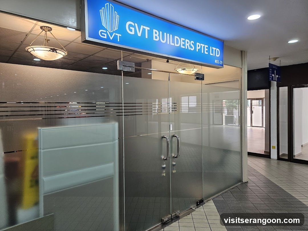 A view of the entrance to GVT Builders Pte Ltd, located at #01-29. The storefront features a large blue sign with white lettering and a logo above glass double doors. The entrance is enclosed by frosted glass panels with horizontal stripes, and the interior appears to be an empty office space.