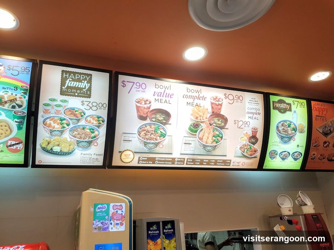 A row of illuminated digital menu boards mounted on an orange ceiling, displaying various meal sets and prices. Below the screens, beverage dispensers and a service counter are partially visible.