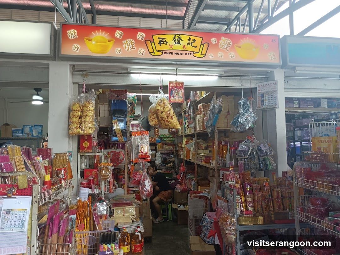 A view of a traditional shop front with a large red and yellow sign reading 'Chye Huat Kee' in English and Chinese characters. The entrance is open, revealing shelves packed with goods. Hanging from the ceiling are bags of snacks and incense sticks. Inside, a person is seated on a stool. Various religious items, candles, and packaged foods are displayed on racks and shelves.
