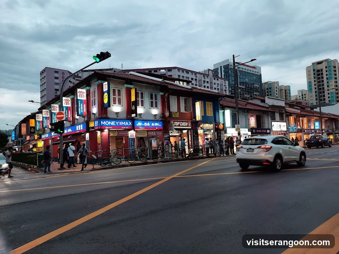 A row of colorful shophouses with various storefronts, including MoneyMax and Top Cash. The street is wet, suggesting recent rain, and features pedestrians and vehicles.