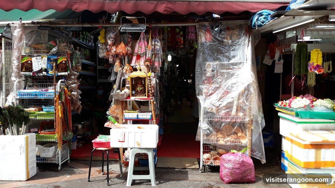 A view of a small market stall or shop front under a red awning. The entrance is covered by clear plastic sheeting. The storefront is densely packed with merchandise, including hanging dried goods, colorful garlands, and packaged items on wire racks. A small shrine sits on a stand near the entrance, and stacks of white and yellow containers are visible on the right.
