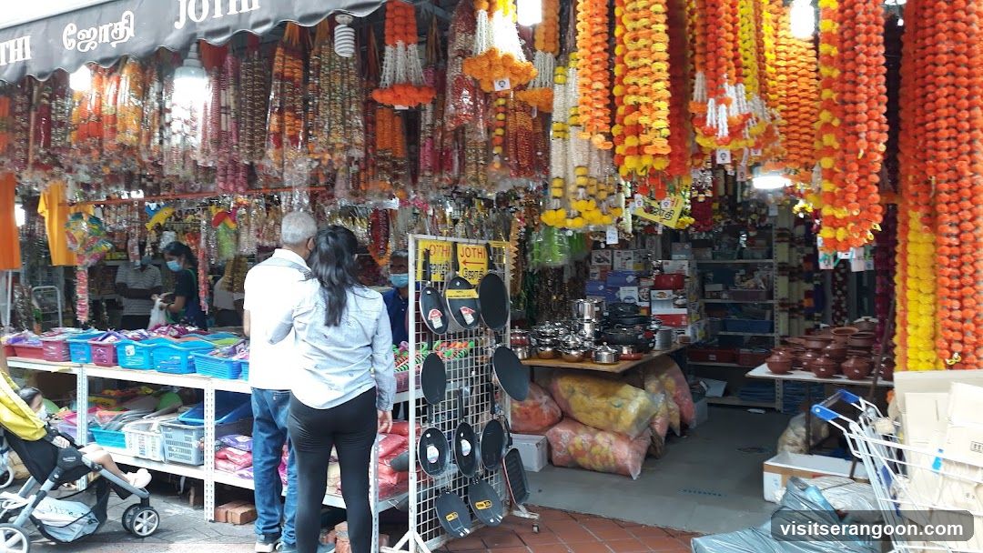 An open-air storefront displaying numerous hanging garlands of orange, yellow, and white flowers, likely marigolds. The shop also sells household items including metal pots, plastic containers, and kitchenware. A sign with the text 'JO THI' is visible on the black awning above the entrance. Two customers are standing at the counter, and a stroller is visible on the left side.