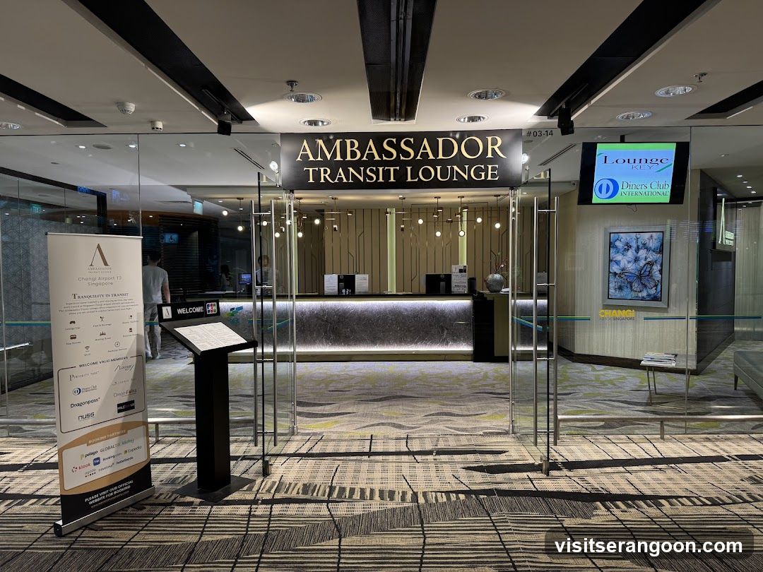A view of the entrance to the Ambassador Transit Lounge, likely within an airport. The image shows glass doors, a reception desk inside, and signage identifying the lounge. A digital screen on the right displays 'Lounge Club' and 'Diners Club International'.