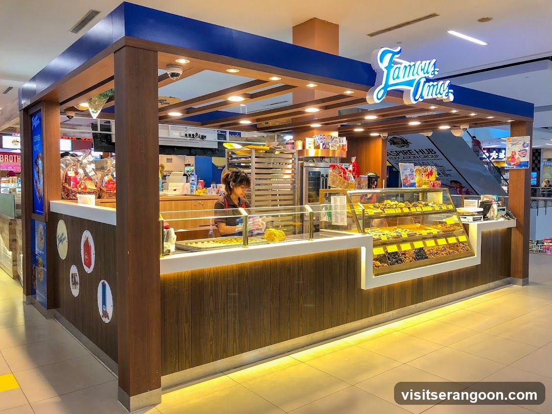 A brightly lit, open-front kiosk located inside a shopping mall. The structure features dark wood paneling, white countertops, and a blue overhead frame. A large, illuminated neon sign reading 'Jamu Jamu' is mounted on the top right. Inside, a staff member is visible behind the counter, and glass display cases showcase various baked goods and pastries. The floor is tiled, and an escalator is visible in the background.
