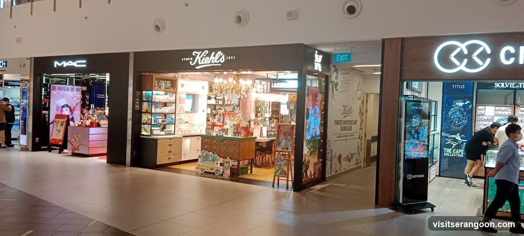 A view of a shopping mall corridor featuring several retail stores. The central store is Kiehl's, identifiable by its white cursive signage above the entrance. To the left is a MAC cosmetics store with illuminated lettering, and to the right is a store with a large infinity symbol logo and the text 'THE CAPE COLLECTION' visible in the window display. People are walking through the mall.