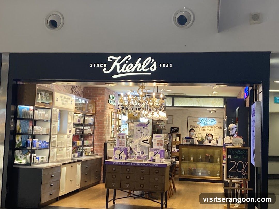 A view of the entrance to a Kiehl's store, identifiable by the large white 'SINCE Kiehl's 1851' logo on the dark blue facade. The interior is visible through the wide opening, featuring a brick wall, glass shelving units stocked with products, and a central display table with purple packaging. Staff members wearing face masks are visible behind a wooden counter in the background.