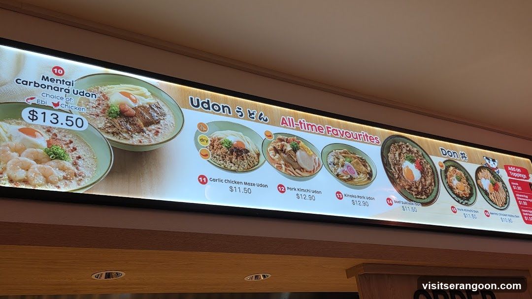 A backlit menu board displaying various noodle dishes, categorized under 'Udon' and 'Donburi'. The board features images of the food, item names, prices, and small icons indicating ingredients like shrimp or chicken.
