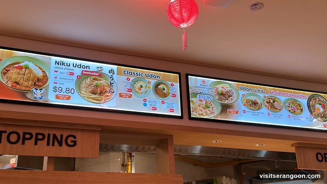 Two illuminated digital menu boards mounted on a wooden wall under a red paper lantern. The left board highlights Niku Udon and Classic Udon options, while the right board displays various All-time Favourites.
