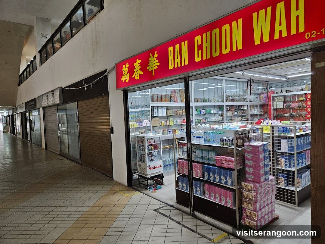 A view of a shop front with a large red sign displaying 'BAN CHOON WAH' in yellow English and Chinese characters. The store has glass doors and windows, revealing shelves stocked with various goods inside. To the left, other storefronts are closed with brown metal shutters down. The floor is tiled, and there is a Yakult refrigerator visible near the entrance.