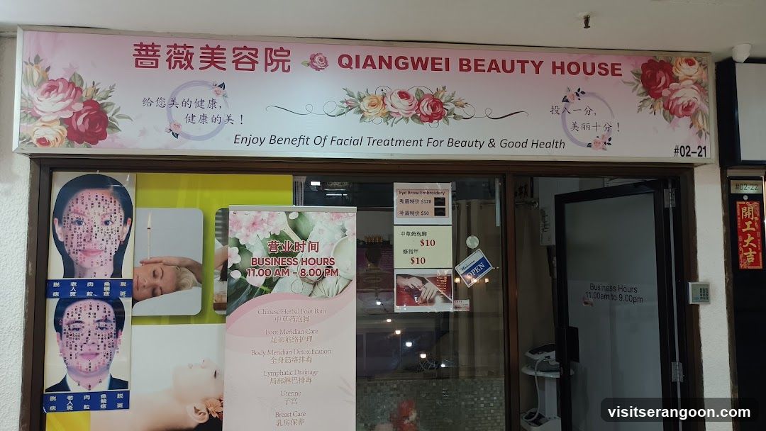 A storefront for a business named 'QIANGWEI BEAUTY HOUSE' with Chinese characters '薔薇美容院' above the entrance. The sign features floral graphics and the slogan 'Enjoy Benefit Of Facial Treatment For Beauty & Good Health'. The entrance has glass doors and windows displaying various posters, including a large vertical banner listing services like Chinese Herbal Foot Bath and Lymphatic Drainage, and smaller signs advertising prices for eye brow threading and eyebrow tinting.
