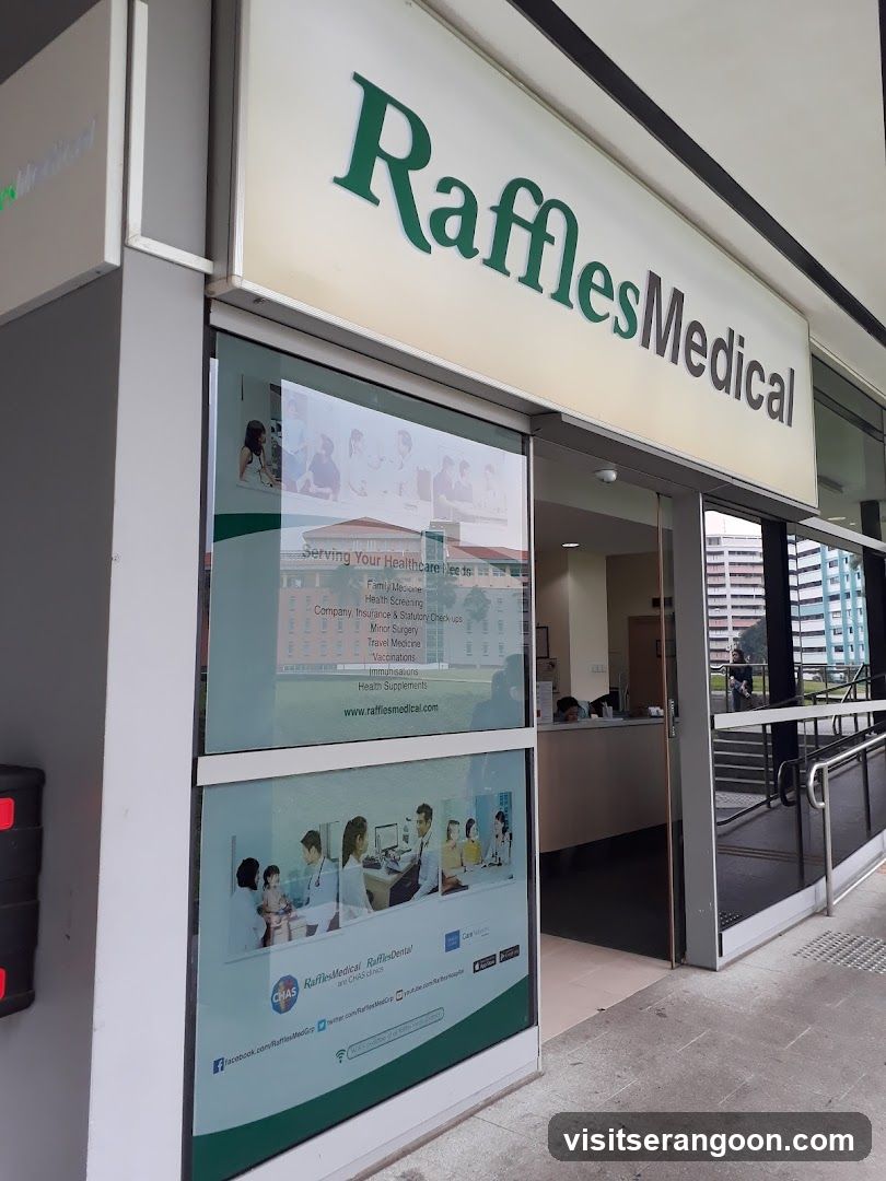 An angled exterior shot of a Raffles Medical clinic entrance. A large cream-colored sign with green and black lettering reading 'Raffles Medical' is mounted above the glass doors. The glass window displays a poster listing services such as Family Medicine, Health Screening, and Minor Surgery, along with the website URL.