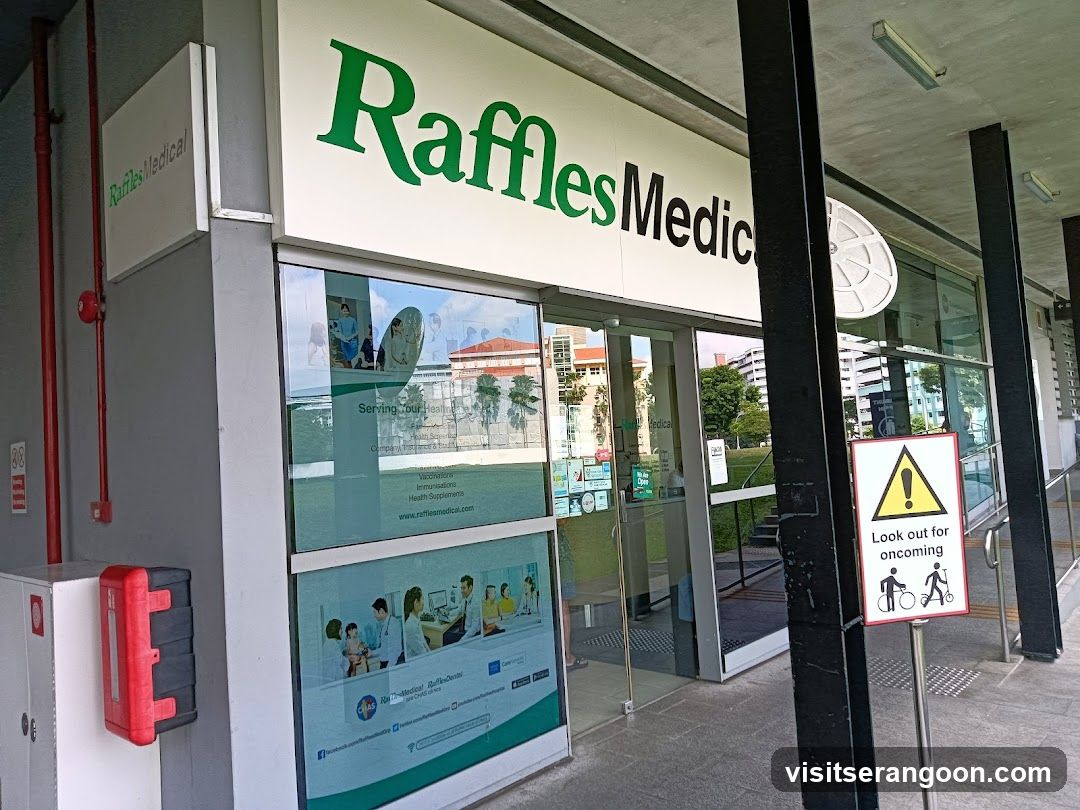 A view of the exterior entrance to a medical clinic. The building features a large white sign with 'Raffles' in green text and 'Medical' in black text. Below the sign, large glass windows display posters with medical imagery and text. To the left, a red fire hose cabinet is mounted on the wall. To the right, a yellow and white safety sign warns about oncoming cyclists and pedestrians. The entrance has glass doors and is situated under a covered walkway.