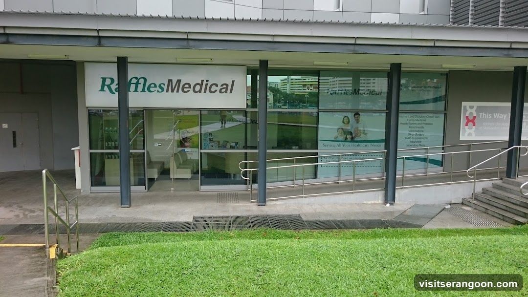 A photo of the exterior storefront of a medical clinic. The building features a white sign with green text reading 'Raffles Medical' above the entrance. The facade includes large glass windows and doors, dark support pillars, and a metal ramp with handrails leading to the entrance. A grassy area is visible in the foreground.