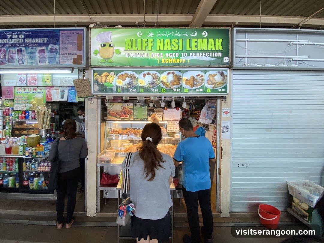 A food stall with a green sign that reads "ALIFF NASI LEMAK BASMATI RICE AGED TO PERFECTION (ASHRAFF)". Below the sign are pictures of different nasi lemak meals with prices. The stall has a display case with food items and people ordering. To the left, there is another stall selling drinks and snacks.