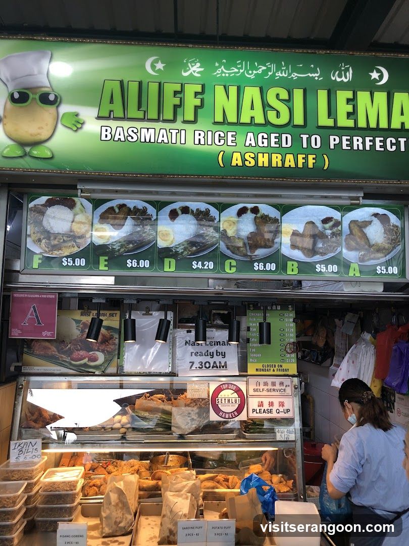 A food stall with a large green sign displaying "ALIFF NASI LEMAK BASMATI RICE AGED TO PERFECTION (ASHRAFF)". Below the sign are images of different nasi lemak sets with prices. The stall has a display case filled with various fried food items and pastries. A sign indicates "Nasi Lemak ready by 7.30AM". Another sign reads "SELF-SERVICE PLEASE Q-UP". A woman in a blue shirt and mask is seen serving customers.