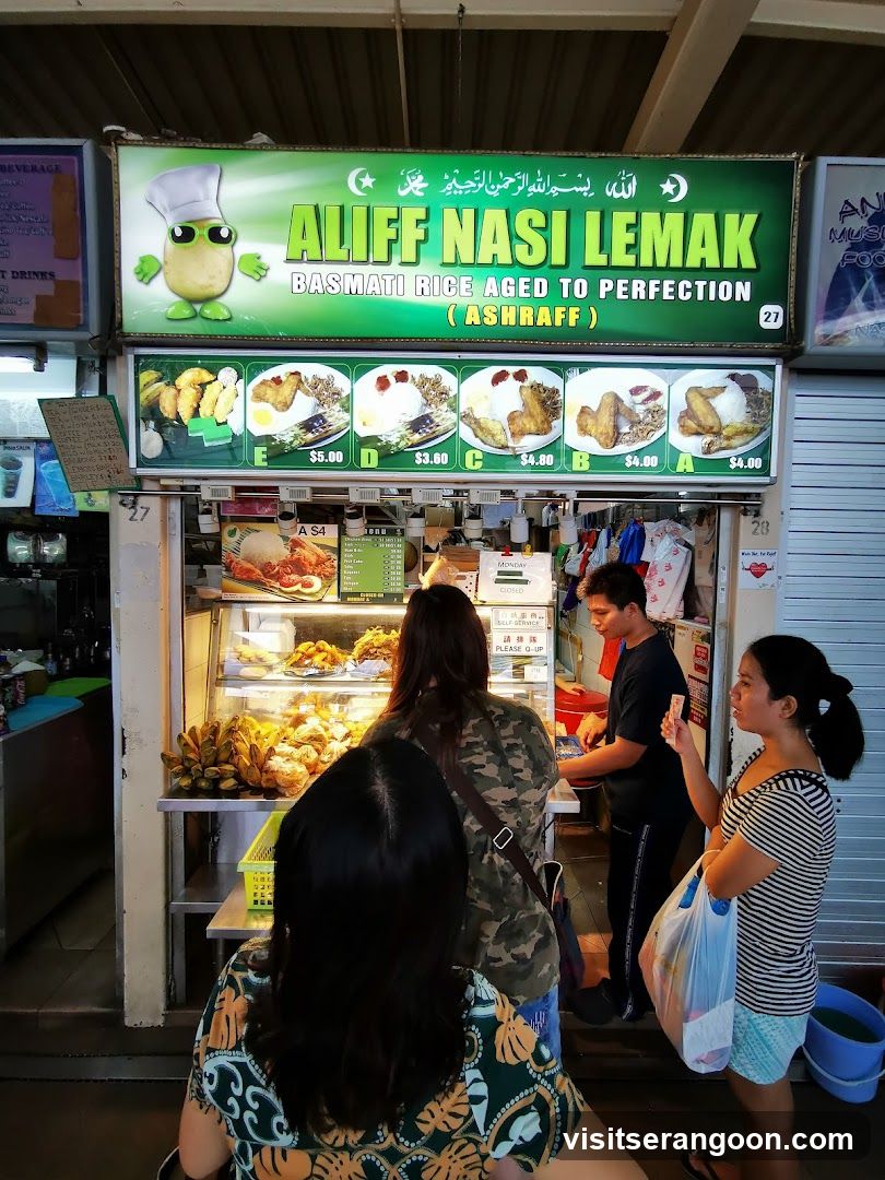 A food stall with a large green sign displaying the business name and images of food items with prices. Below the sign is a display case filled with various fried foods and a bunch of bananas. Several customers are visible, some ordering and others waiting.