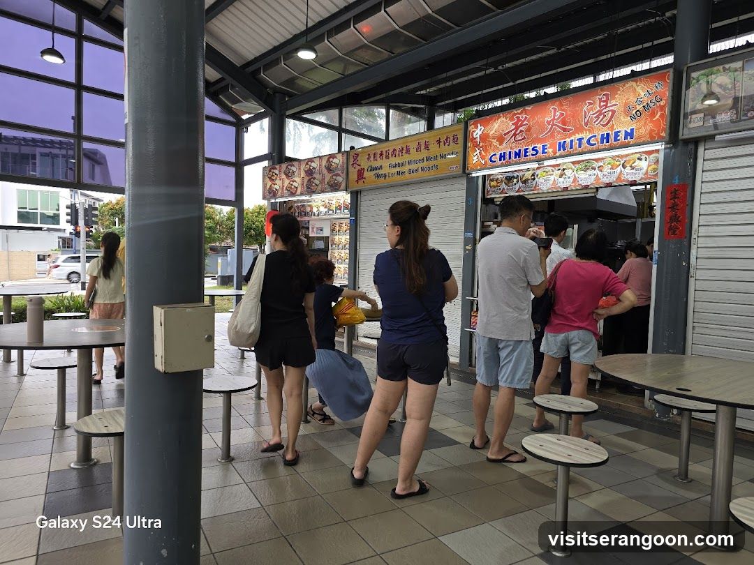 A food stall with a large sign that reads "CHINESE KITCHEN" in English and Chinese characters. Below the main sign, there are smaller signs with pictures of food and prices. Several people are standing in line to order. The stall appears to be part of an outdoor food court or hawker center, with tables and chairs visible in the foreground.