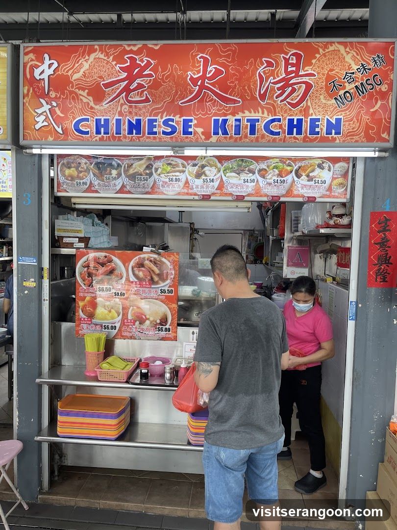 A food stall with a large sign that says "CHINESE KITCHEN" in English and Chinese characters. Below the main sign, there are pictures of various soup dishes with their prices. Two people are standing at the counter, one ordering and the other serving. The stall appears to be in an outdoor food court or market.