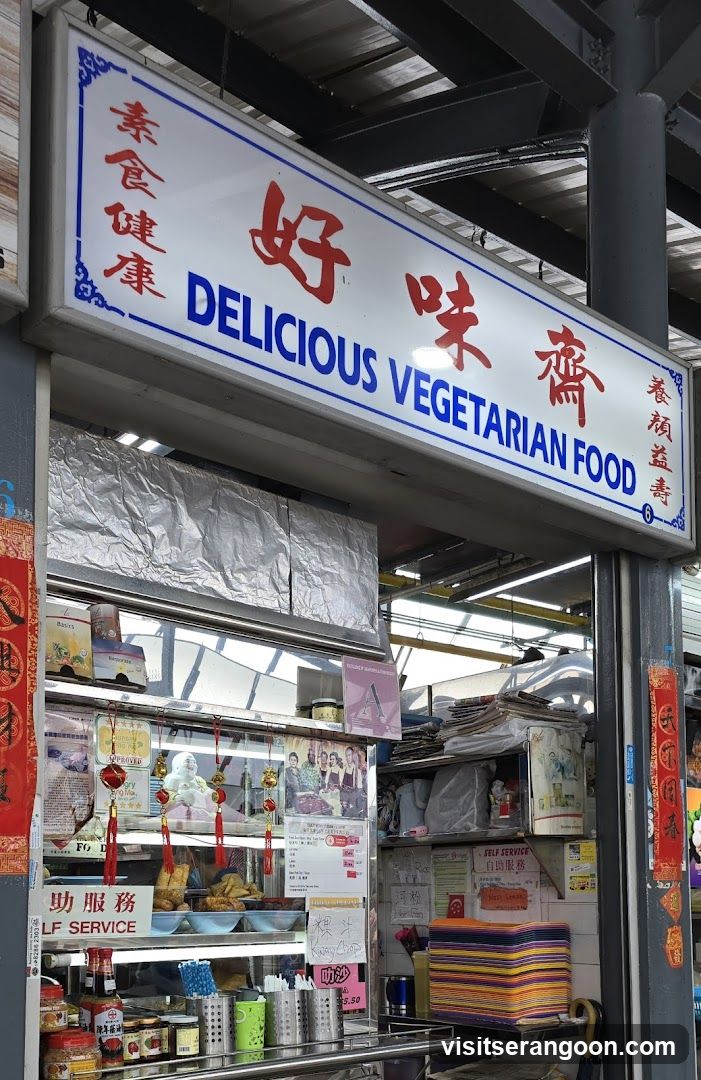 A stall with a prominent sign reading "DELICIOUS VEGETARIAN FOOD" in English and Chinese characters. Below the sign, the stall displays various food items, condiments, and promotional materials. There are also stacks of colorful plates and a self-service sign.
